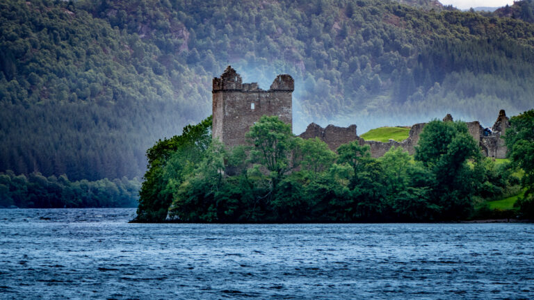 A view of Loch Ness Urquhart castle Ruins, with rich green trees Infront of the castle and a calm mist behind the castle.