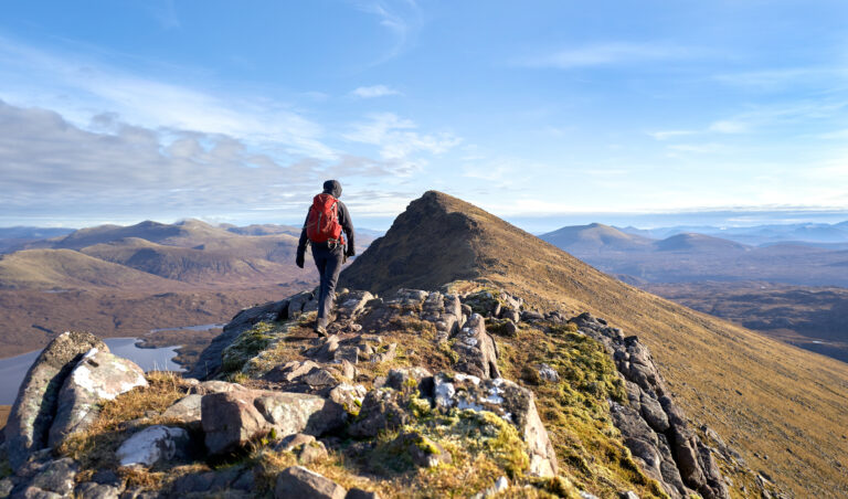 A hiker walking towards the summit of Sgurr an Tuill Bhain along a narrow rocky ridge in the Scottish Highlands on a sunny winters day.