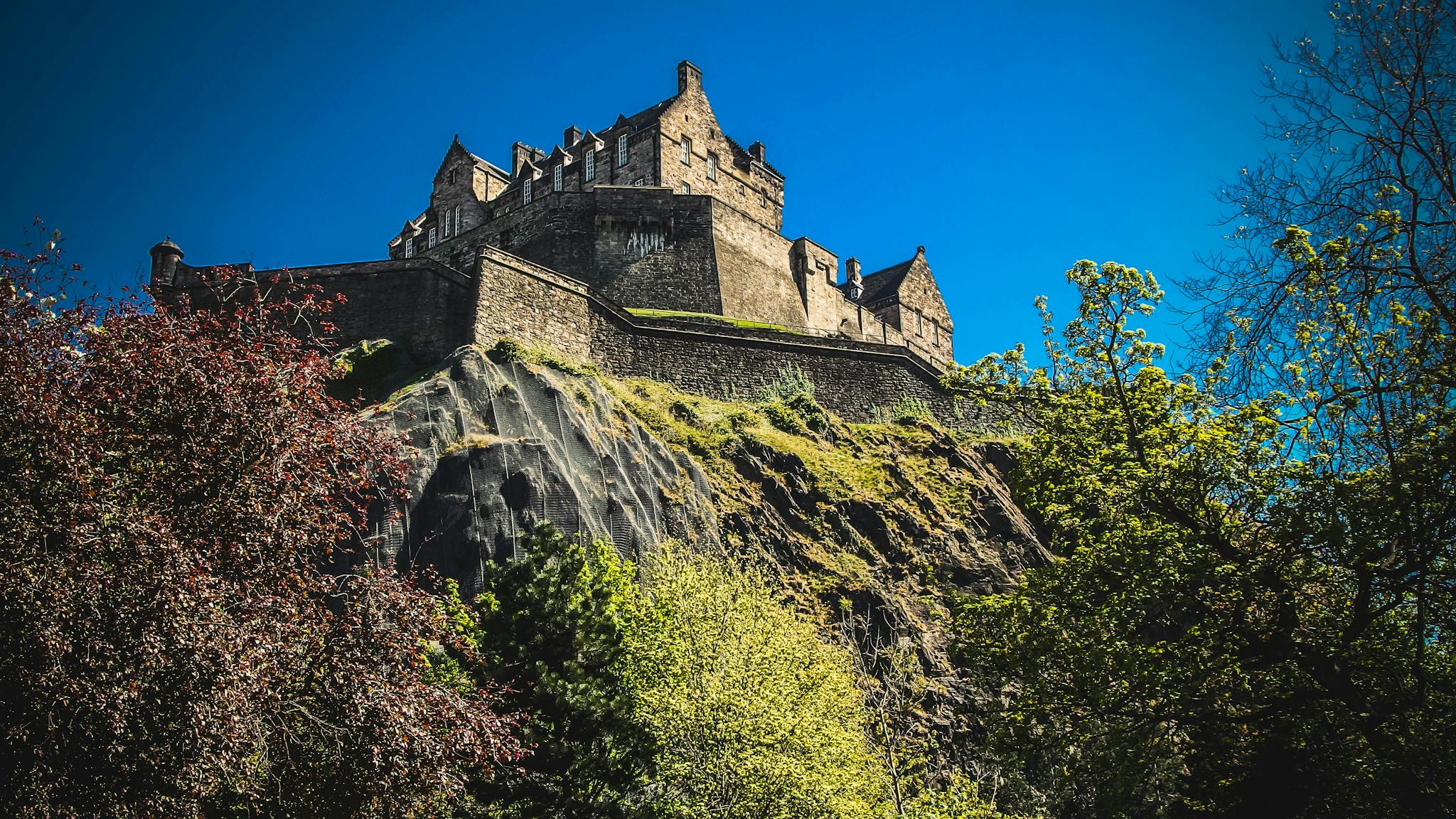 Iconic Edinburgh Castle atop Castle Rock under a clear blue sky, surrounded by lush greenery.