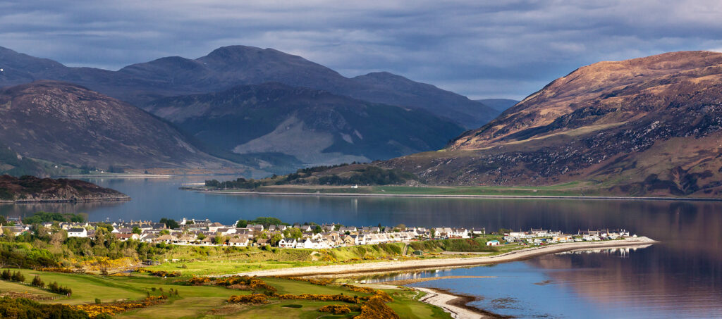 A view of the town of Ullapool with sun shining upon the town and surrounding water.